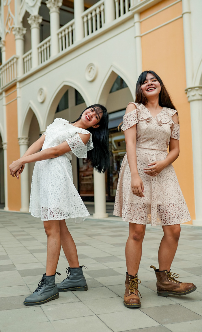 Two women smiling and posing in stylish lace dresses outdoors during the day.