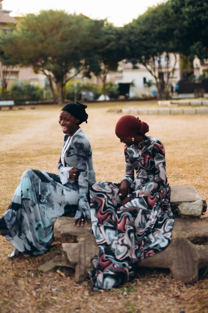 Two women in colorful dresses sitting on stone outdoors, sharing a joyful moment in a park setting.