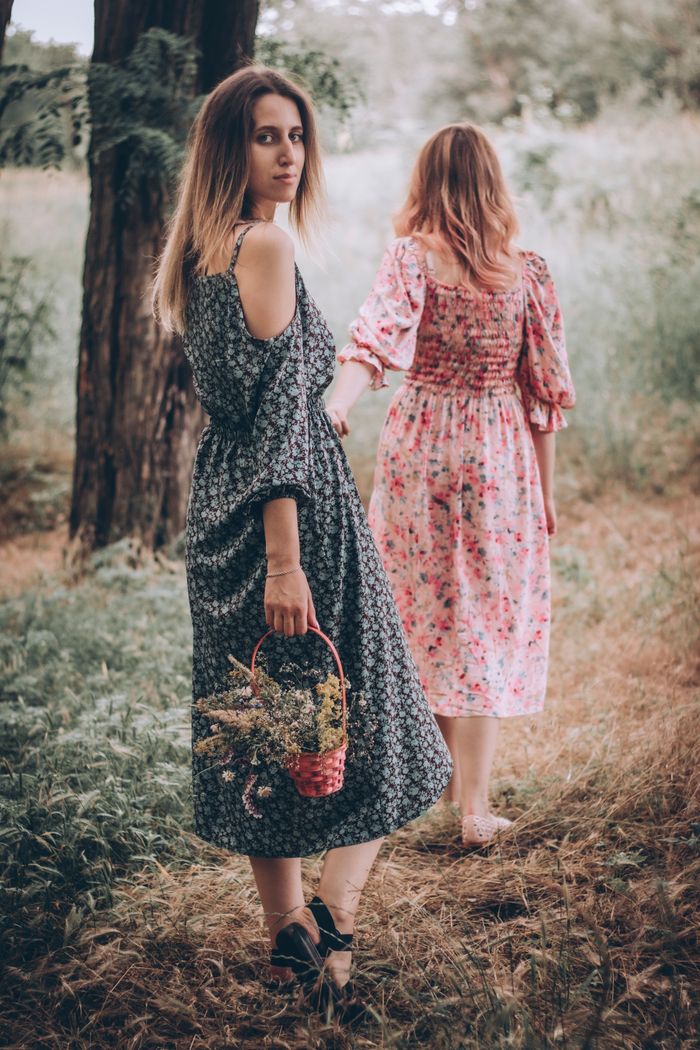 Two women in floral dresses holding basket, walking in a serene forest setting.